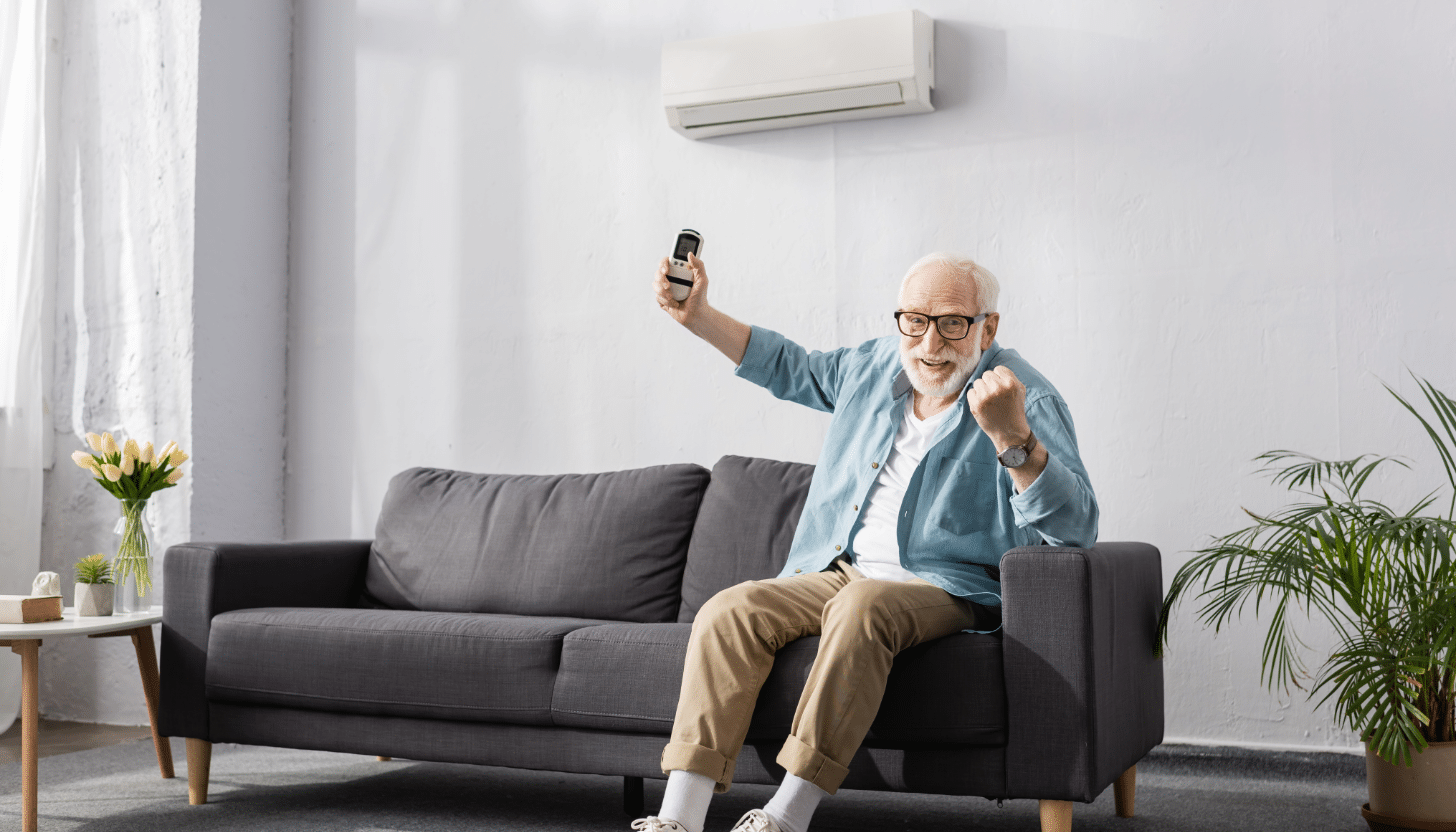 Elderly man sitting on a dark gray couch, smiling and raising his fist in excitement while holding a remote control for his multi-zone hvac system. A multi-zone AC unit is mounted on the white wall above, providing efficient cooling. The bright living room features modern decor, highlighting the comfort and convenience of a best multi-zone mini split system.